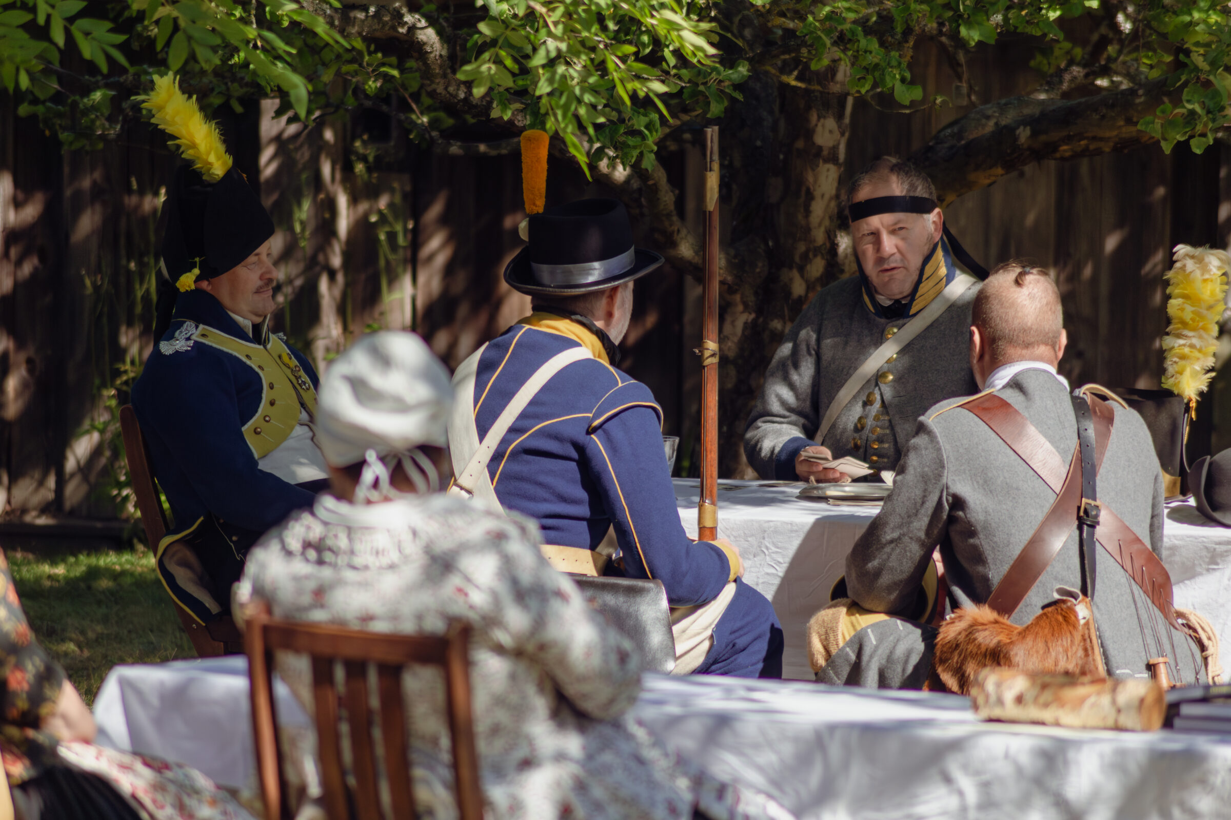 Personer i historiska militärkläder spelar kort i skuggan av ett träd.